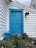 A vibrant blue door is set against white horizontal siding, with the number 12 beside it. A dark outdoor light fixture is mounted above the number, adding a rustic touch. The bottom of the frame is filled with lush greenery and blooming pink flowers in pots, adding a pop of color and freshness.