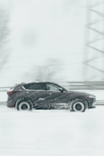 A dark SUV is driving through a snowy landscape, with snow falling heavily, creating a blur effect. The vehicle moves swiftly on a road that is barely visible through the dense snow. Trees and structures are faintly visible in the background.