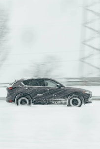 A dark SUV is driving through a snowy landscape, with snow falling heavily, creating a blur effect. The vehicle moves swiftly on a road that is barely visible through the dense snow. Trees and structures are faintly visible in the background.
