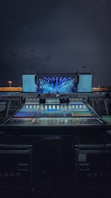 An outdoor stage setup is illuminated with vibrant blue and purple lights, with a large sound mixing console in the foreground. The sky is dark, indicating nighttime, and there are two large screens on either side of the stage.