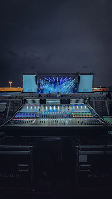 An outdoor stage setup is illuminated with vibrant blue and purple lights, with a large sound mixing console in the foreground. The sky is dark, indicating nighttime, and there are two large screens on either side of the stage.