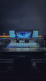 An outdoor stage setup is illuminated with vibrant blue and purple lights, with a large sound mixing console in the foreground. The sky is dark, indicating nighttime, and there are two large screens on either side of the stage.