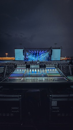 An outdoor stage setup is illuminated with vibrant blue and purple lights, with a large sound mixing console in the foreground. The sky is dark, indicating nighttime, and there are two large screens on either side of the stage.