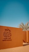 A reddish-brown wall with an emblem and the text 'Liwa Village' in both English and Arabic. Palm trees rise in the background under a clear blue sky, with a sandy foreground.