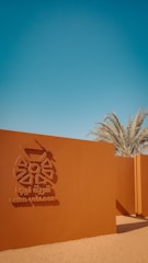 A reddish-brown wall with an emblem and the text 'Liwa Village' in both English and Arabic. Palm trees rise in the background under a clear blue sky, with a sandy foreground.