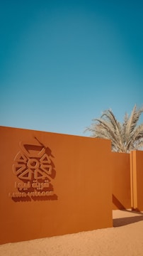 A reddish-brown wall with an emblem and the text 'Liwa Village' in both English and Arabic. Palm trees rise in the background under a clear blue sky, with a sandy foreground.