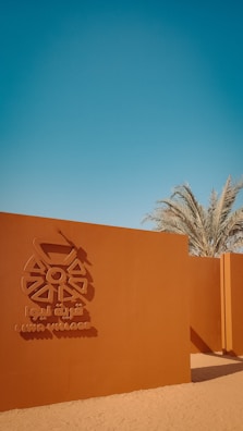 A reddish-brown wall with an emblem and the text 'Liwa Village' in both English and Arabic. Palm trees rise in the background under a clear blue sky, with a sandy foreground.
