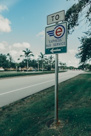 A signpost is situated by the side of a roadway, indicating an express bus service route. The signpost features a blue and white sign with an emblem of an 'e' and a wing. Palm trees and lush greenery line the way, with a clear blue sky above. The road appears to be relatively empty, suggesting a quiet moment.