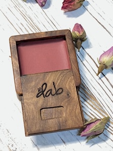 A wooden compact case containing a pink cosmetic product. The surface of the case is engraved with the word 'dab'. Around the case, there are dried rosebuds scattered on a white wooden background with visible grain lines.