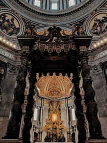 An intricately designed church interior with a grand altar featuring ornate sculptures and golden decorations. The architecture includes large, twisted columns supporting an elaborately adorned canopy. The ceiling displays detailed artwork with religious imagery and Latin inscriptions. Light filters in through the windows, illuminating the Baroque style decor.