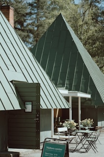 A modern visitors center with distinct triangular roofs made of green metal sheets is surrounded by trees. The foreground includes a sign mentioning the visitors center and several sets of outdoor tables and chairs with potted plants nearby, creating an inviting atmosphere.