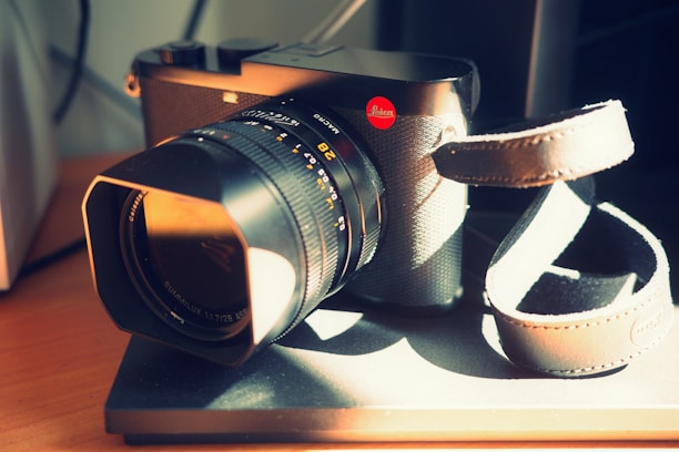 A high-end camera with a large lens sits on a flat surface. The camera body features a textured finish and a prominent red logo on the side. A leather strap is attached to the camera, draping over the surface. The lighting creates a warm and inviting ambiance, with shadows adding depth to the scene.