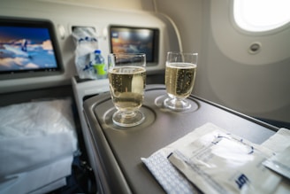 Two glasses filled with a bubbly drink sit on an aircraft tray table, accompanied by packaged snacks. The cabin appears to be well-lit, and two screens in the background display content related to aviation.