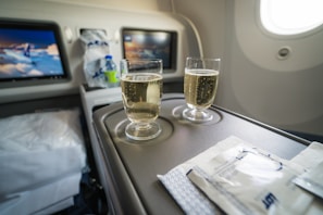 Two glasses filled with a bubbly drink sit on an aircraft tray table, accompanied by packaged snacks. The cabin appears to be well-lit, and two screens in the background display content related to aviation.