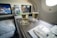 Two glasses filled with a bubbly drink sit on an aircraft tray table, accompanied by packaged snacks. The cabin appears to be well-lit, and two screens in the background display content related to aviation.
