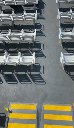 Rows of white benches casting distinct shadows on a gray, textured surface. The benches are arranged in orderly lines, suggesting a seating area. Bright yellow non-slip strips mark steps at the bottom of the image.