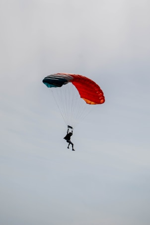 A silhouette of a person is parachuting against a cloudy sky. The parachute is vibrantly colored with red, orange, and teal sections, creating a striking contrast against the overcast background.