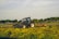 A tractor with the brand name 'Valtra' is operating in a field with green vegetation in the foreground. The background features a rural scene with houses and trees under a partly cloudy sky.