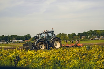 A tractor with the brand name 'Valtra' is operating in a field with green vegetation in the foreground. The background features a rural scene with houses and trees under a partly cloudy sky.