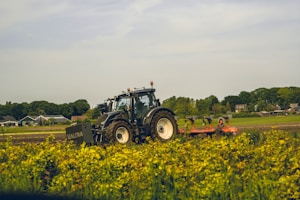 A tractor with the brand name 'Valtra' is operating in a field with green vegetation in the foreground. The background features a rural scene with houses and trees under a partly cloudy sky.