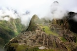 Ancient stone structures are nestled within a mountainous landscape, surrounded by lush greenery and partially obscured by clouds. The site features terraces and buildings constructed from large stone blocks, showcasing intricate and historical architecture.