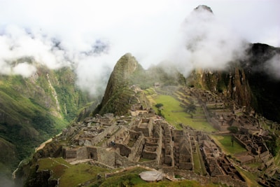 Ancient stone structures are nestled within a mountainous landscape, surrounded by lush greenery and partially obscured by clouds. The site features terraces and buildings constructed from large stone blocks, showcasing intricate and historical architecture.