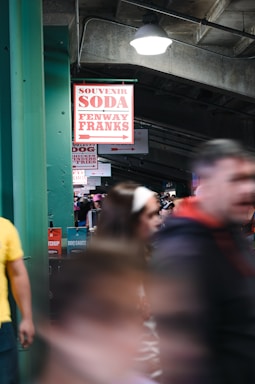 A crowded indoor area featuring a concession sign displaying options such as souvenirs, soda, and Fenway Franks. The scene is dynamic, with people moving past in the foreground. The setting appears to be a stadium or event venue, indicated by the signs and bustling atmosphere.