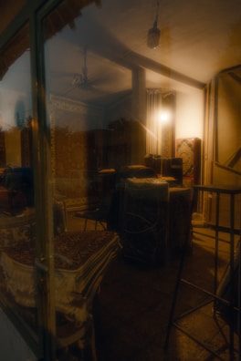 A dimly lit interior with stacked carpets and rugs visible through a glass window. The room is illuminated by a soft, warm light, highlighting intricate patterns on the fabrics. The scene has a reflective surface, possibly due to the glass, adding a hazy effect. Stools and a ceiling fan are present in the background.