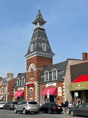A historical fire department building is depicted with a tall, pointed tower topped with a weather vane. The structure is made of red brick with stone accents around the windows and doors. Bright red awnings are above the entrances, which are labeled as 'Hose No 1' and 'Engine No 1'. Several cars are parked along the street, and a few pedestrians are visible. The sky is clear and blue, suggesting a sunny day.
