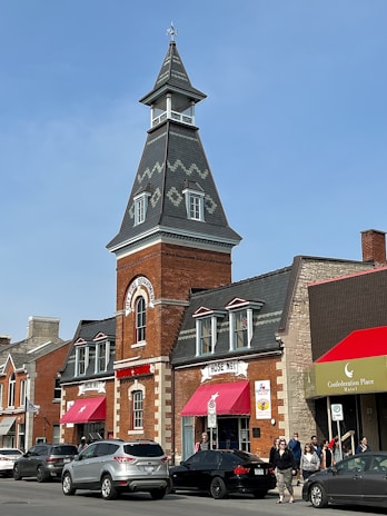 A historical fire department building is depicted with a tall, pointed tower topped with a weather vane. The structure is made of red brick with stone accents around the windows and doors. Bright red awnings are above the entrances, which are labeled as 'Hose No 1' and 'Engine No 1'. Several cars are parked along the street, and a few pedestrians are visible. The sky is clear and blue, suggesting a sunny day.