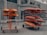 Two racks of kayaks are parked on a gravel surface outside a building. The rack on the left holds orange school kayaks labeled 'KAYAKS SCOLAIRES'. The rack on the right has a mix of red and gray kayaks, labeled 'KAYAKS CADRES'. Both racks are mounted on wheels for easy mobility. The building in the background appears industrial with large windows and a plain facade.