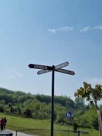 A metal signpost with multiple directional arrows pointing towards various locations, including Zenica and Spomenici Smehn. It is set against a background of green, lush hills under a clear blue sky. A small tree is present on the right, and a pedestrian sign is visible below the signpost. Two people are seen walking on the road toward the left side of the image.