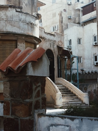 A vintage architectural setting featuring an old stone building with arched windows and a curved staircase leading up to an entrance. The building exudes a weathered charm with its textured stonework, surrounded by other residential structures displaying a similar aged aesthetic.