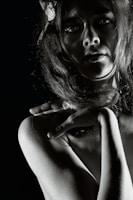 A dramatic black and white portrait of a person with long hair, partially illuminated, creating strong shadows across the face and body. The hand is delicately placed on the shoulder, adding an element of grace and intrigue. The overall composition highlights the contours and textures in a striking manner.