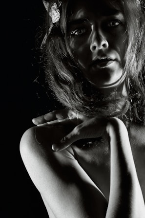 A dramatic black and white portrait of a person with long hair, partially illuminated, creating strong shadows across the face and body. The hand is delicately placed on the shoulder, adding an element of grace and intrigue. The overall composition highlights the contours and textures in a striking manner.