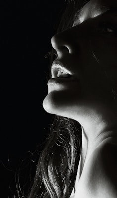 A close-up profile of a person's lower face and neck illuminated dramatically from a low angle, creating deep shadows and highlights. The light accentuates the lips, chin, and part of the nose, with hair framing the face.