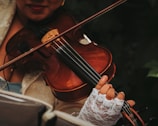 A person is playing a violin, with a focus on the fingers pressing the strings. The musician is wearing lace gloves and part of the instrument's body and bow are visible. The background is dark and out of focus, highlighting the rich, warm tones of the violin.