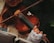 A person is playing a violin, with a focus on the fingers pressing the strings. The musician is wearing lace gloves and part of the instrument's body and bow are visible. The background is dark and out of focus, highlighting the rich, warm tones of the violin.