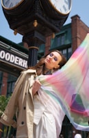 A woman wearing a beige trench coat gracefully poses with a colorful, flowing scarf in front of a vintage black clock with Roman numerals. The background features old brick buildings and a sign with partially visible letters.
