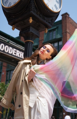 A woman wearing a beige trench coat gracefully poses with a colorful, flowing scarf in front of a vintage black clock with Roman numerals. The background features old brick buildings and a sign with partially visible letters.