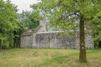 A rustic stone building with a slate roof is nestled among lush green trees. The grass around the building is neatly trimmed, and the scene conveys tranquility and isolation.