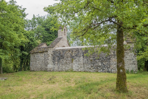 A rustic stone building with a slate roof is nestled among lush green trees. The grass around the building is neatly trimmed, and the scene conveys tranquility and isolation.