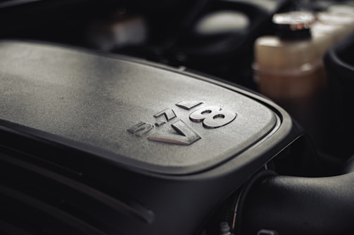 A close-up view of a 5.7L V8 engine cover with bold lettering. The metallic surface has a textured finish, and the background shows parts of the engine compartment.