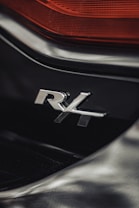 A close-up view of a vehicle's badge featuring the letters 'R4' in chrome, set against a sleek black surface. Above the badge, a section of red tail light is visible, adding a contrast to the overall dark tones.