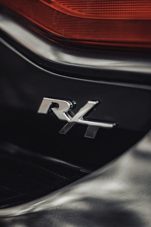 A close-up view of a vehicle's badge featuring the letters 'R4' in chrome, set against a sleek black surface. Above the badge, a section of red tail light is visible, adding a contrast to the overall dark tones.