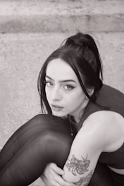 A person with long dark hair tied in a ponytail is sitting with their knees slightly raised. They have a nose piercing and winged eyeliner. A tattoo is visible on the arm featuring an intricate design. The background appears to be a textured surface, possibly concrete.
