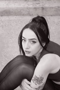 A person with long dark hair tied in a ponytail is sitting with their knees slightly raised. They have a nose piercing and winged eyeliner. A tattoo is visible on the arm featuring an intricate design. The background appears to be a textured surface, possibly concrete.