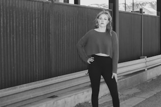 A woman stands confidently with one hand on her hip, wearing a cropped sweater and leggings. She is positioned next to a metal fence and guardrail, with mountains visible in the background.