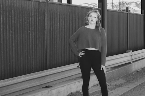 A woman stands confidently with one hand on her hip, wearing a cropped sweater and leggings. She is positioned next to a metal fence and guardrail, with mountains visible in the background.