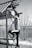 A person in athletic attire is climbing a metal structure outdoors. The background consists of bare trees, suggesting a winter or early spring setting. The individual is wearing a cap and sports shoes, indicating they might be engaging in an outdoor exercise or adventure activity.